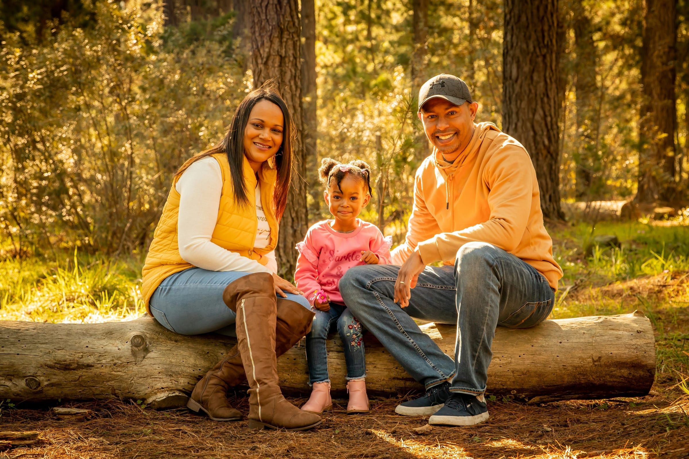 A family of three sitting together on a fallen log in a sunlit pine forest.